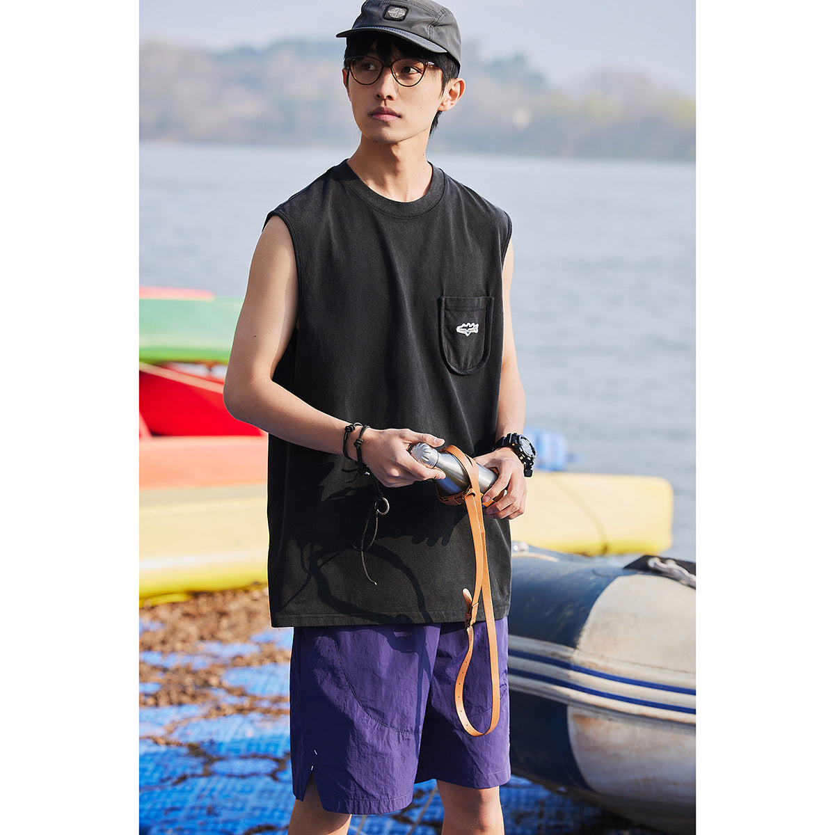 Model wearing a loose black sleeveless shirt with a chest pocket and purple shorts by the water.