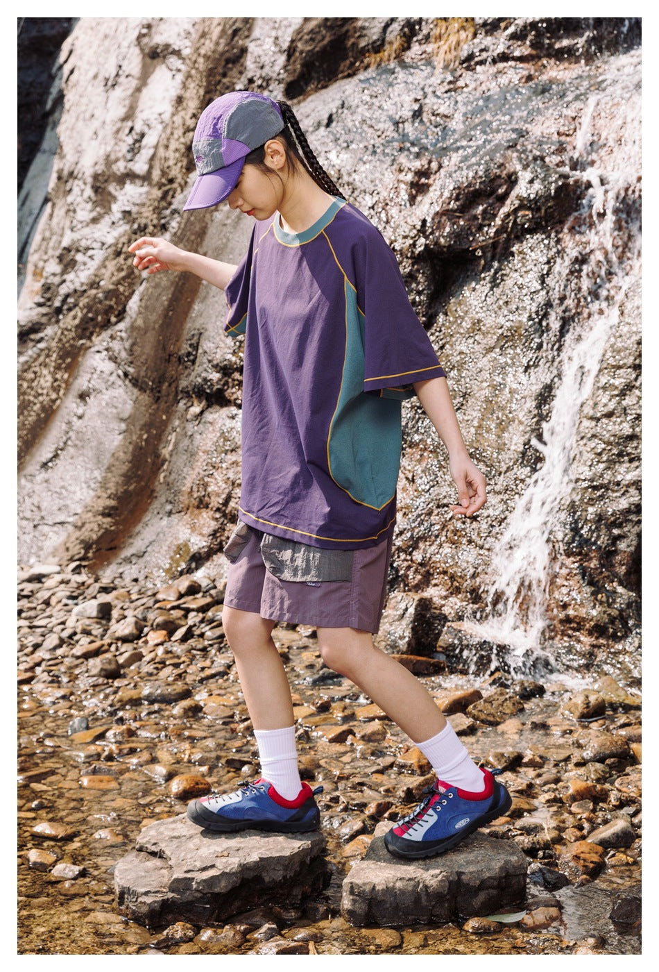 Side view of a model stepping across rocks in the purple T-shirt with teal side panels, shorts and a cap