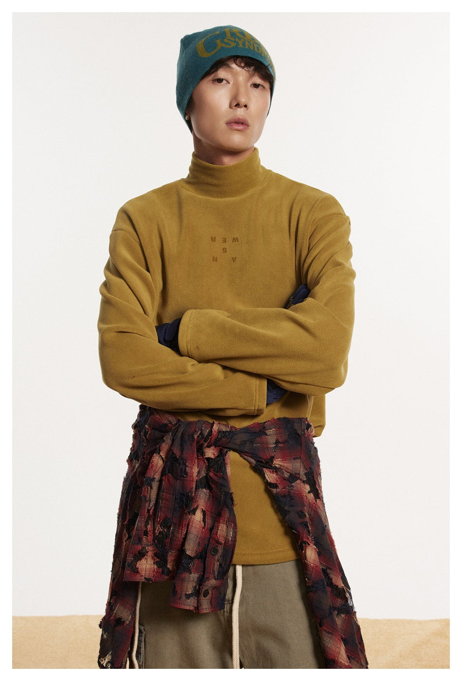 Model wearing a mustard high-neck top with small chest lettering, shown with a teal beanie and arms crossed.
