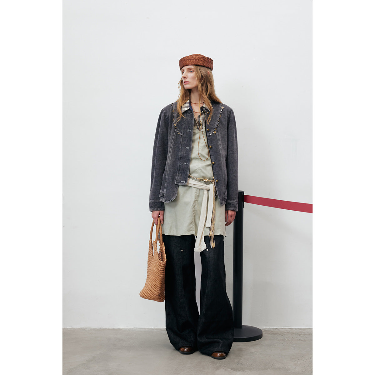 Model wearing a charcoal embellished denim jacket over a pale tunic and wide-leg black trousers, with a woven brown bag.