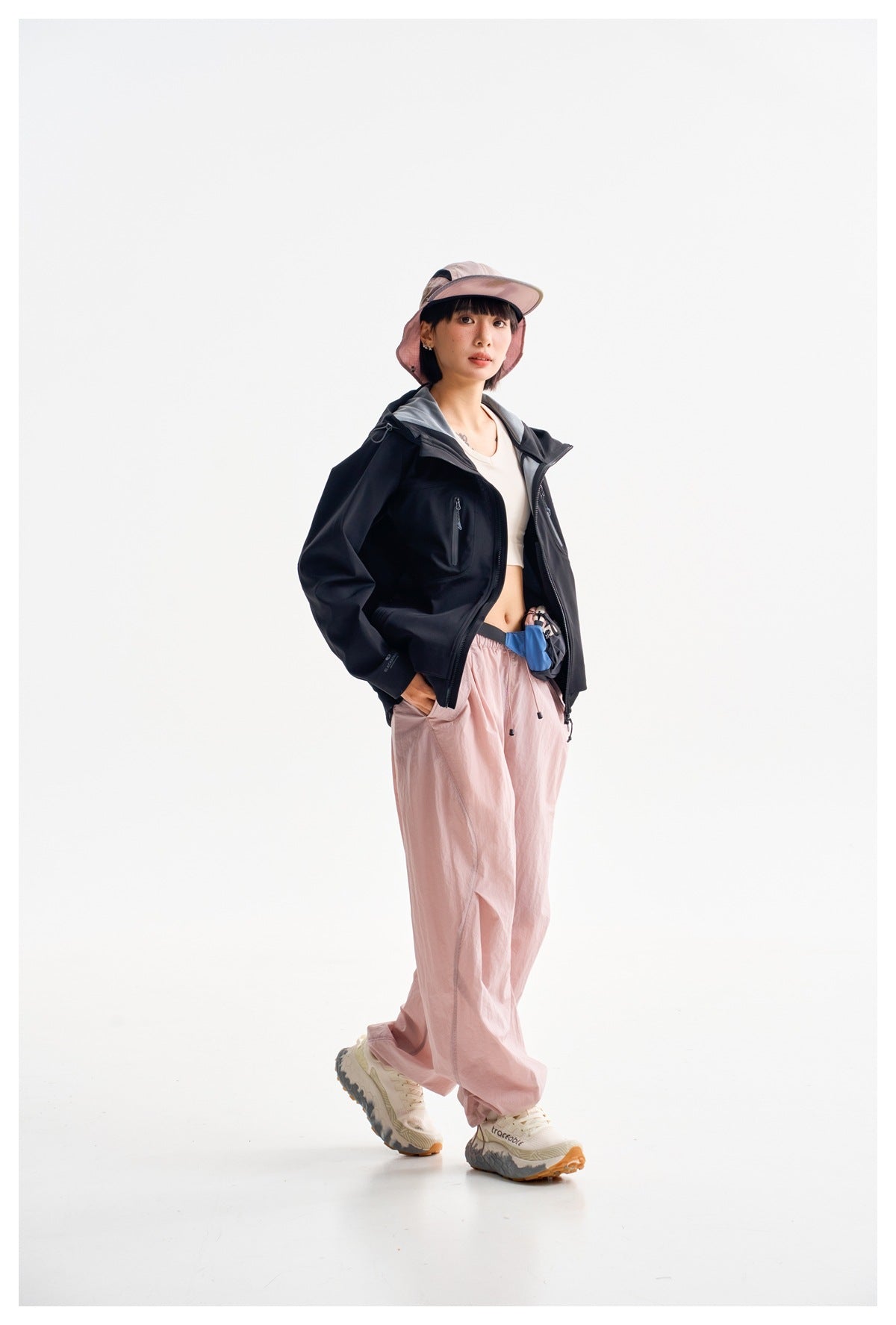 Model wearing an open black hooded jacket with a zip chest pocket, pink trousers, and a pink sun hat.