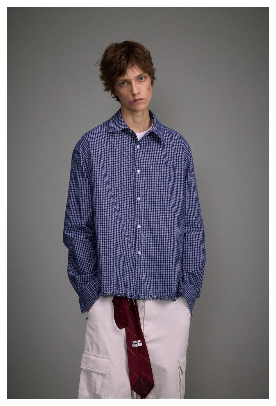 Model wearing a blue-and-white checked button-up shirt with a chest pocket and frayed hem.