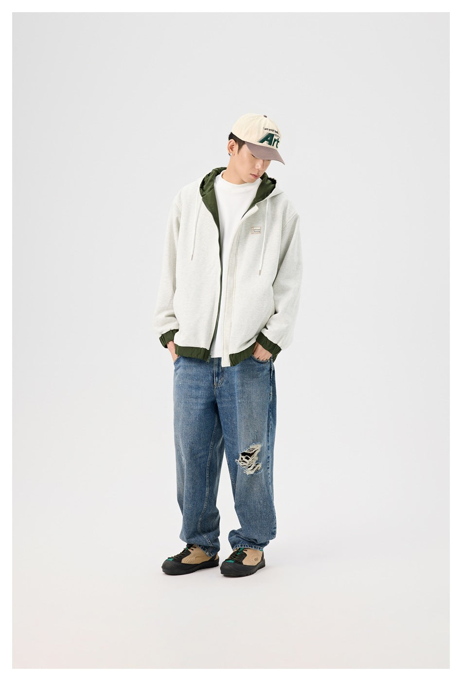Model wearing a light gray zip hoodie with dark green hood lining, cuffs and hem, styled with jeans and a cap.