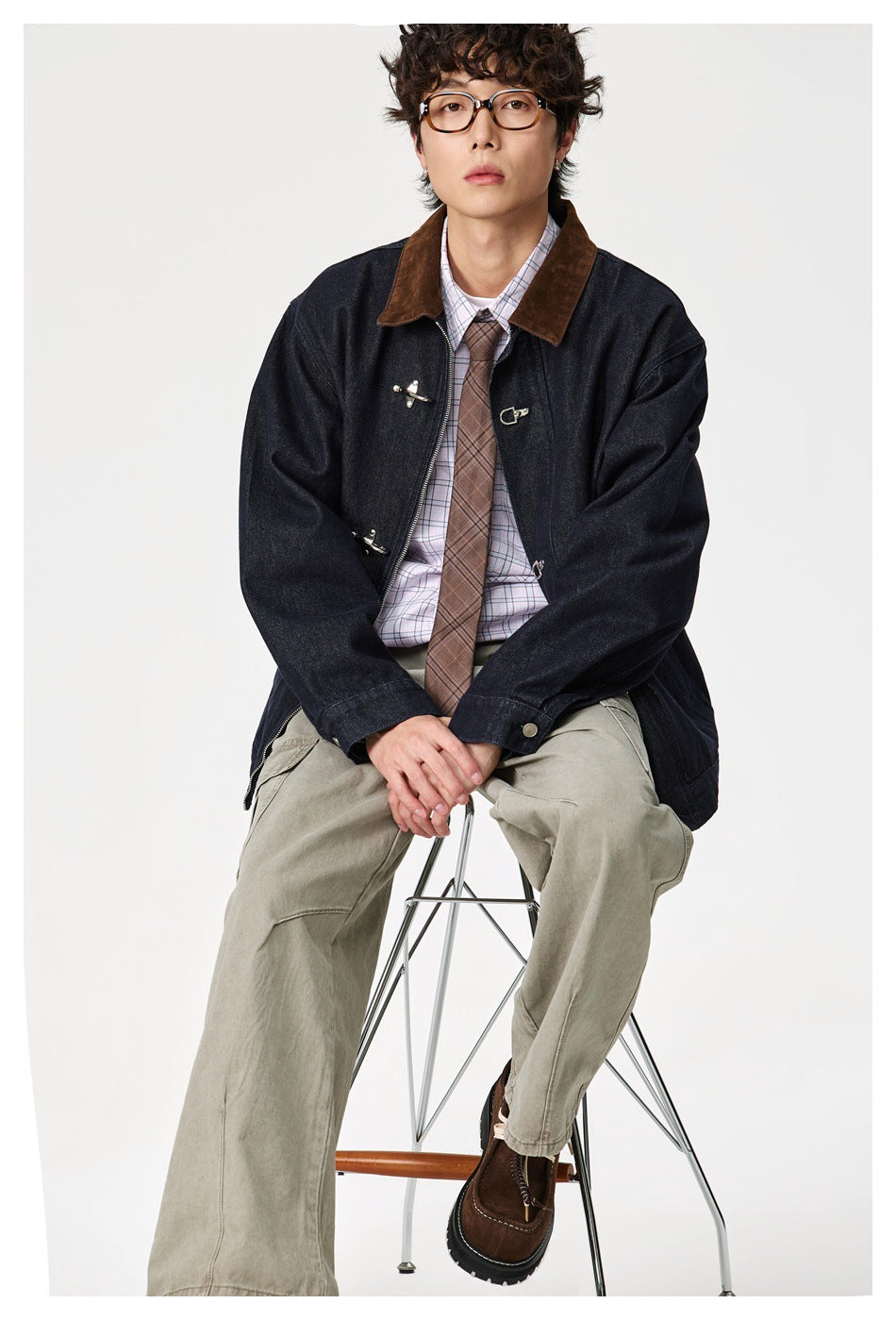 Seated model in navy denim jacket with hook clasps and brown collar, styled with plaid shirt and olive pants