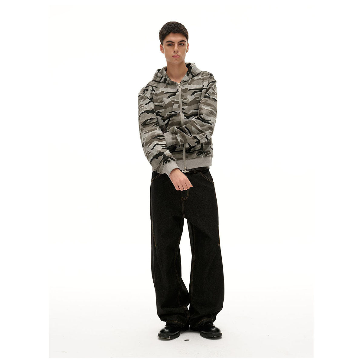 Model wearing a gray camouflage zip-up hoodie with the hood down and loose black jeans, shown from the front.