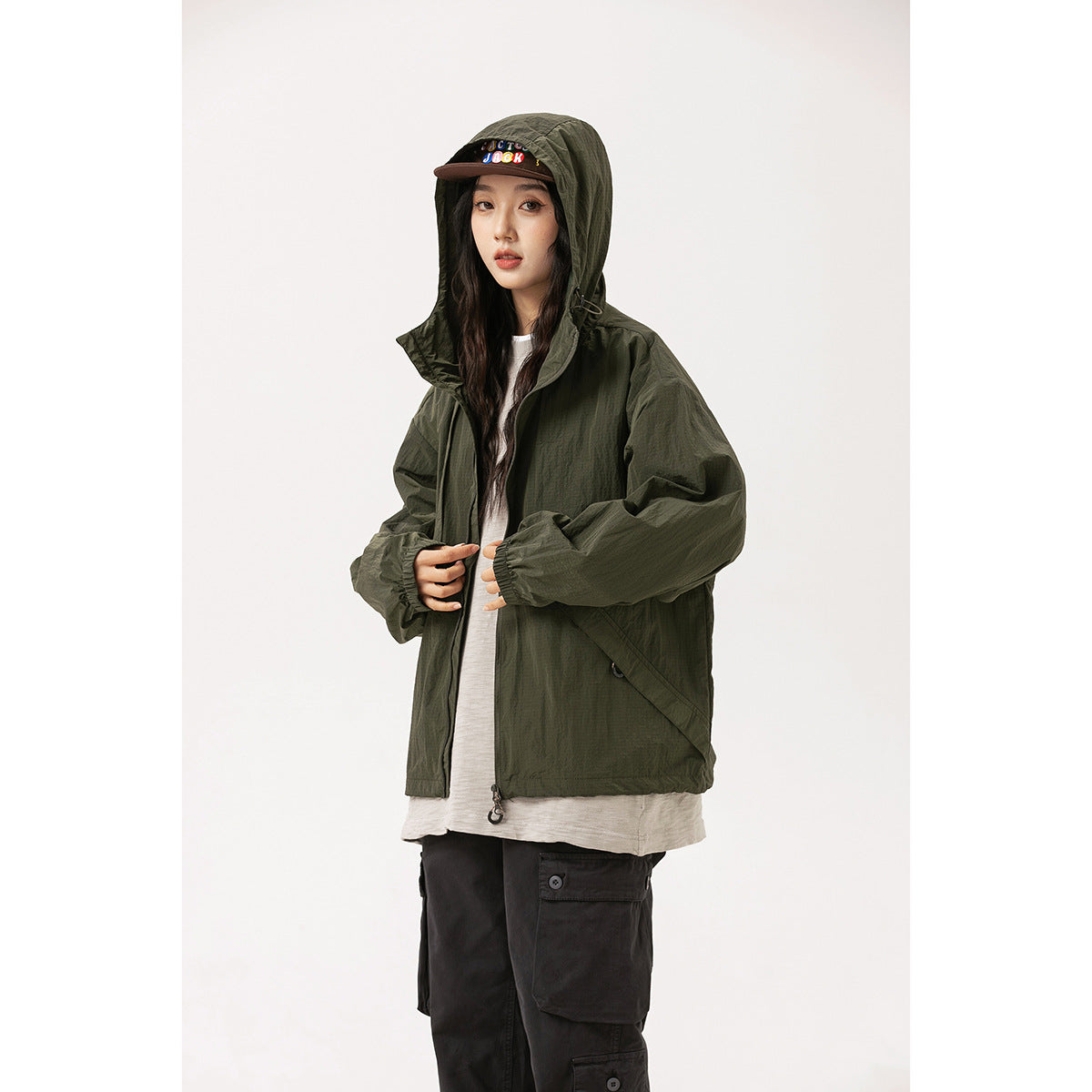Model wearing an olive hooded jacket with a front zip and large pockets; colorful lettering is visible on the cap.