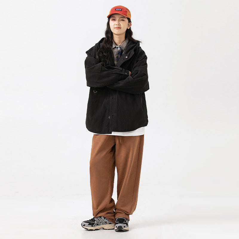 Model wearing a black hooded jacket over a plaid shirt, with loose brown pants and an orange cap.