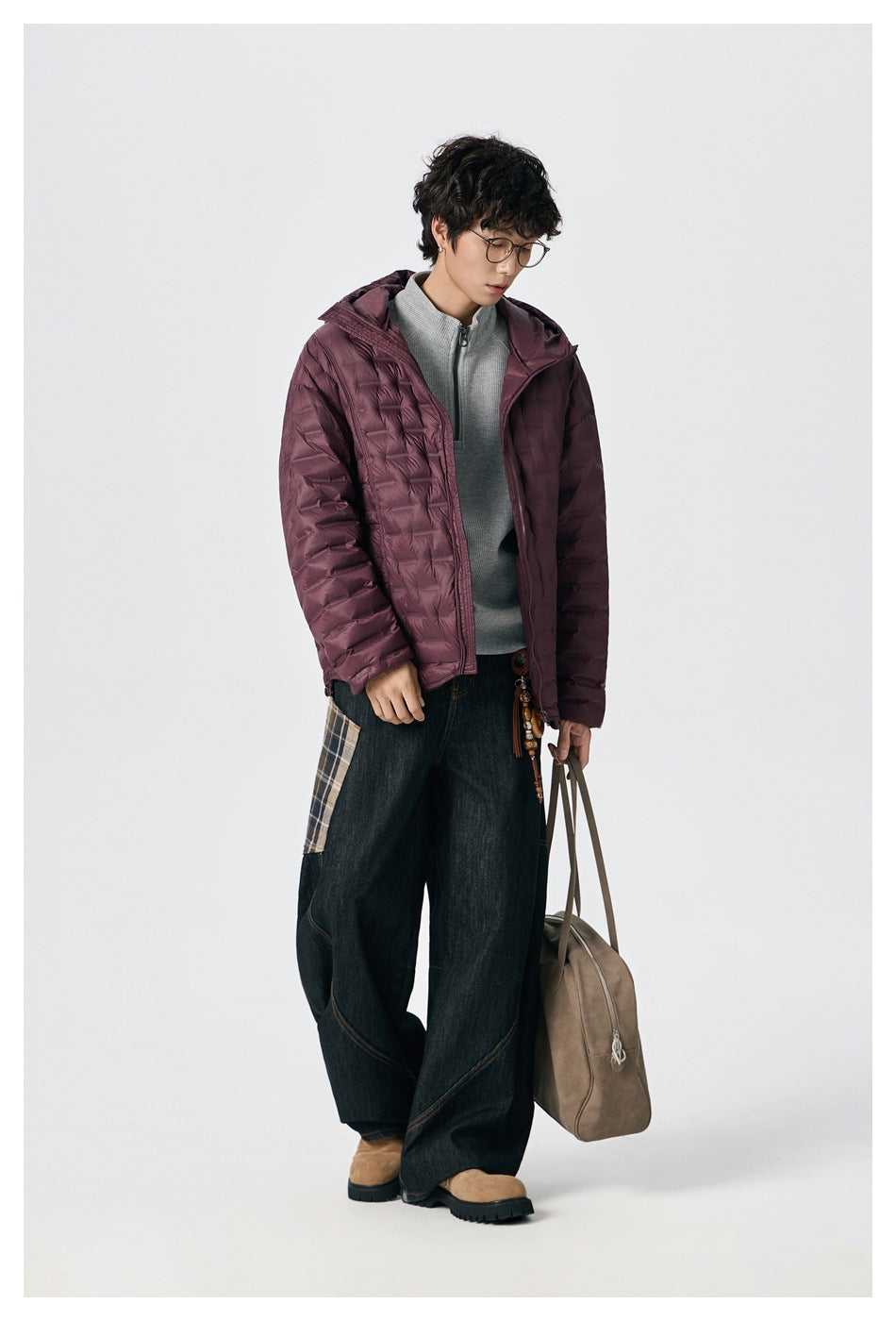 Model wearing a burgundy hooded quilted jacket and dark loose jeans, standing at an angle with a bag.