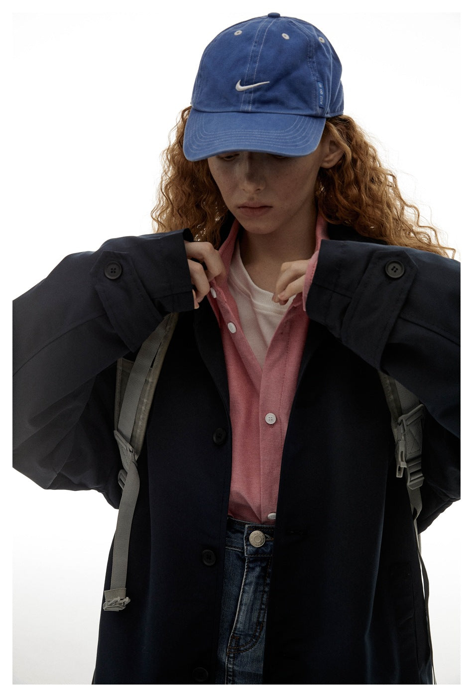 Close view of a model in a dark navy coat, pink shirt, blue cap and backpack straps.