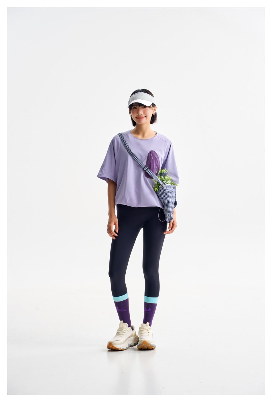 Model wearing a lavender short-sleeve top, dark leggings, purple socks, and a gingham crossbody bag.