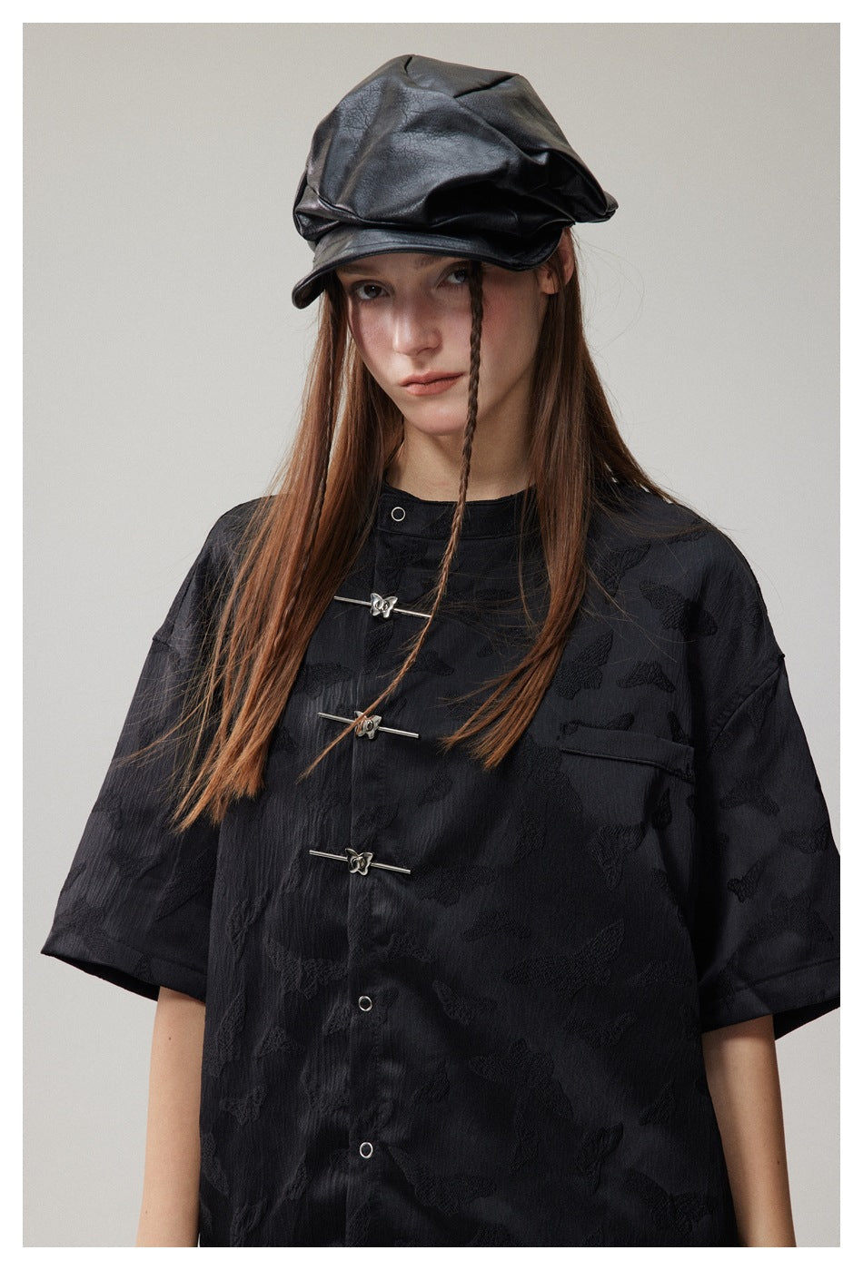 Close view of a black butterfly-patterned shirt with silver frog closures, modeled with a black cap.