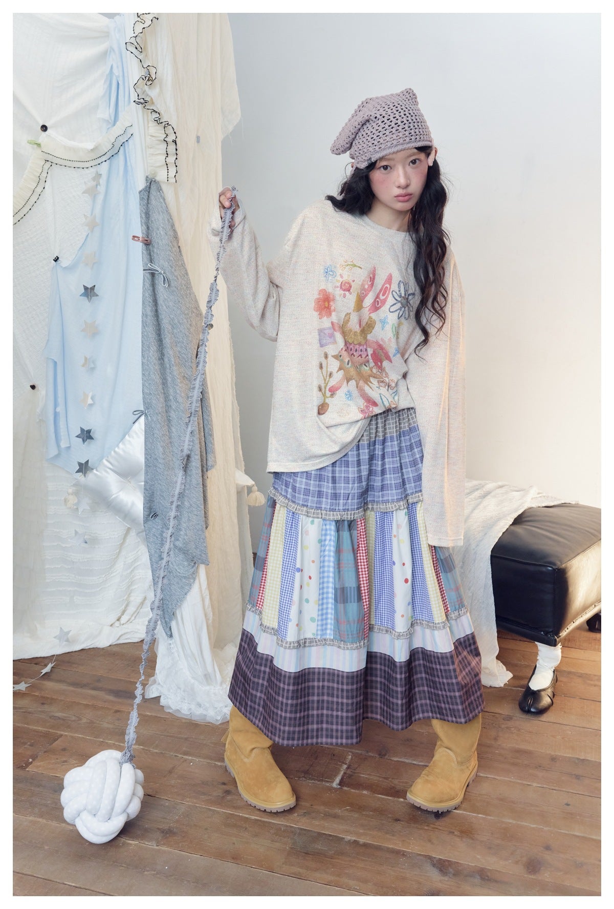 Model wearing a cream long-sleeve printed top oversized with a patchwork plaid skirt, knit headscarf and tan boots