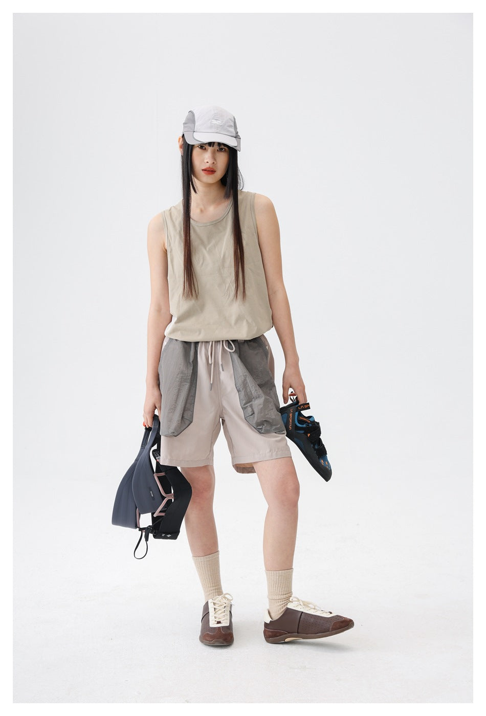 Model wearing a sand beige sleeveless tank top with gray shorts and a cap, holding climbing shoes and a harness
