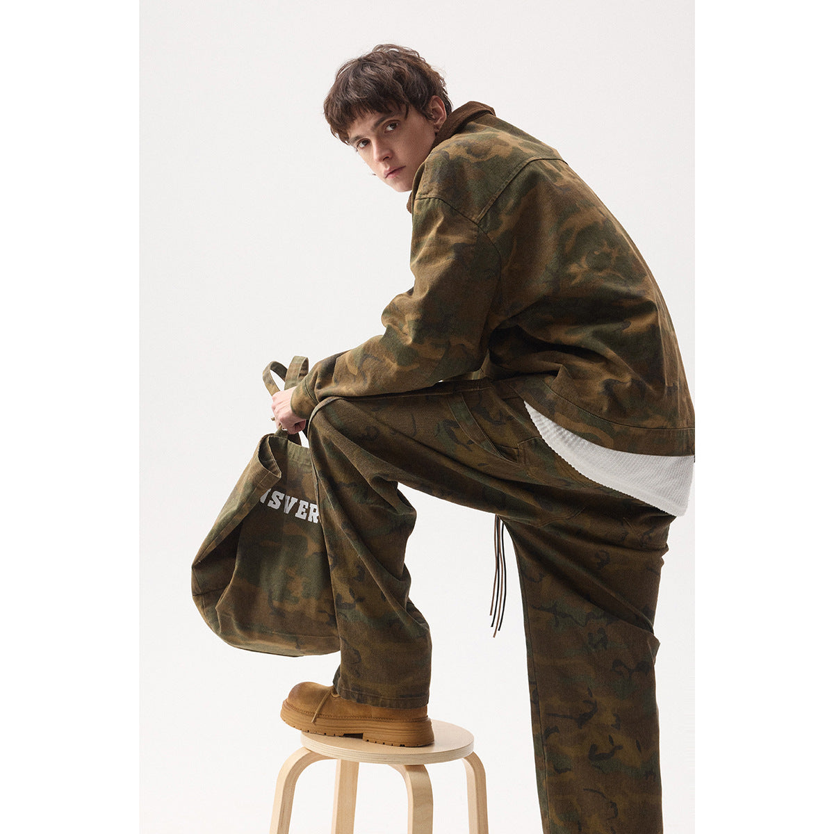 Model crouching in a brown-green camouflage jacket and loose trousers, showing the jacket’s back and side while holding a