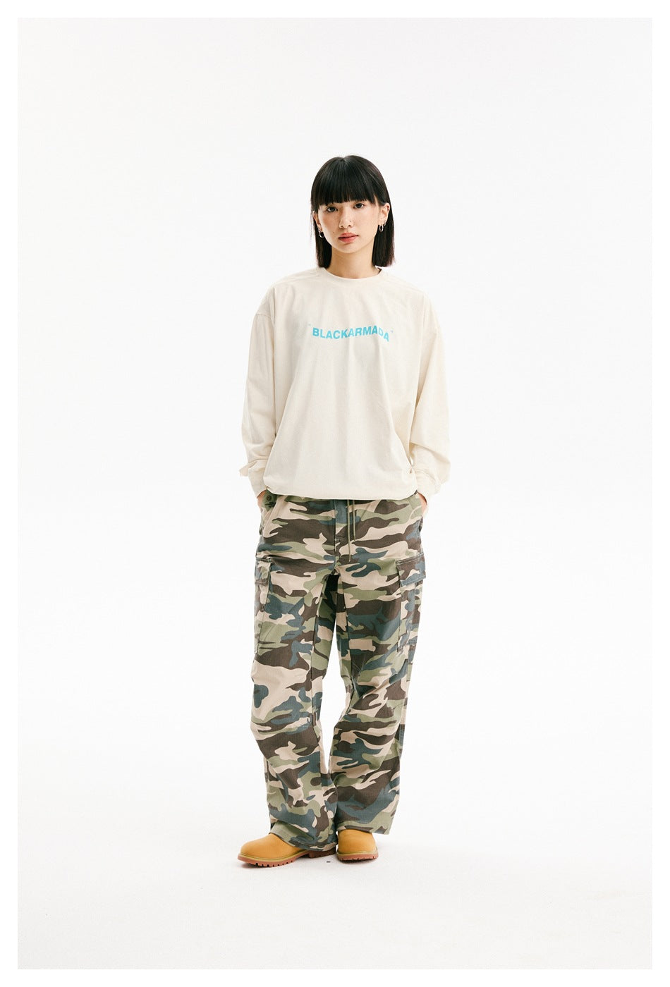 Front view of a model in an ivory long-sleeve shirt with turquoise chest lettering and loose camouflage cargo pants.