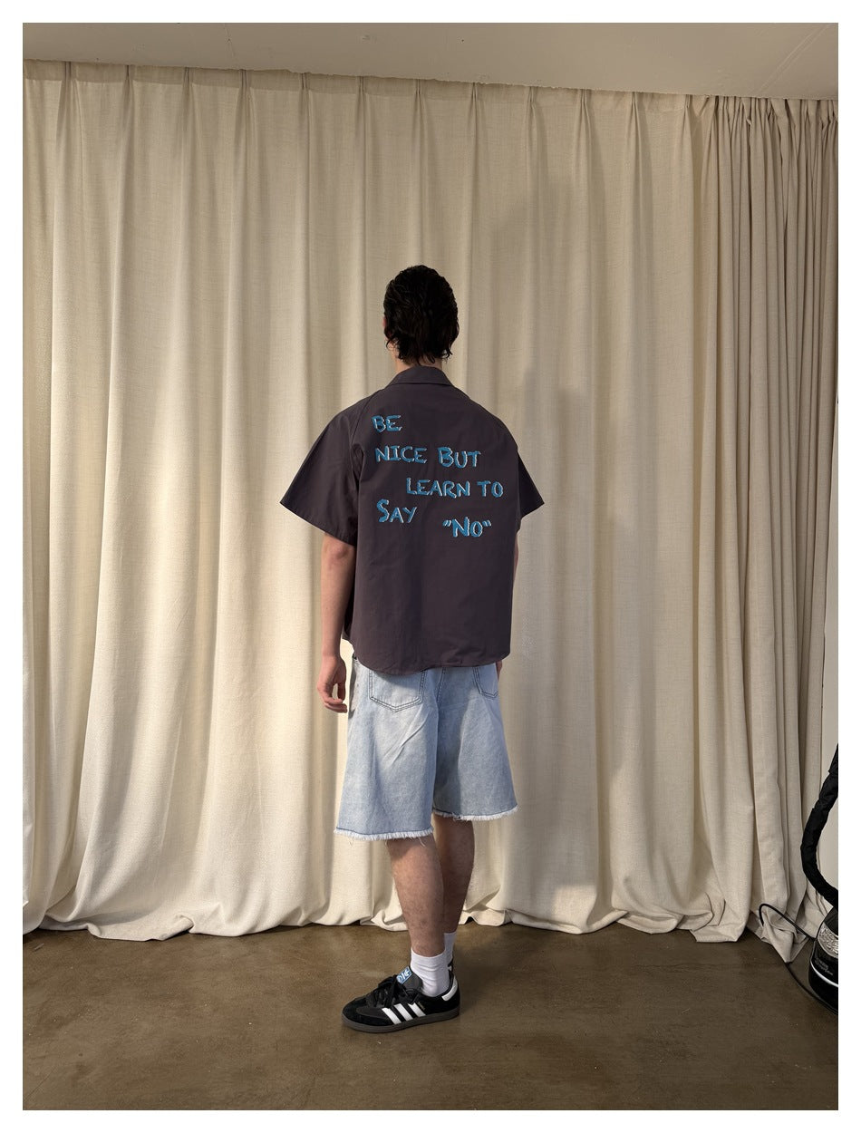 Rear view of a model wearing a charcoal short-sleeve shirt with blue lettering across the back and light denim shorts.