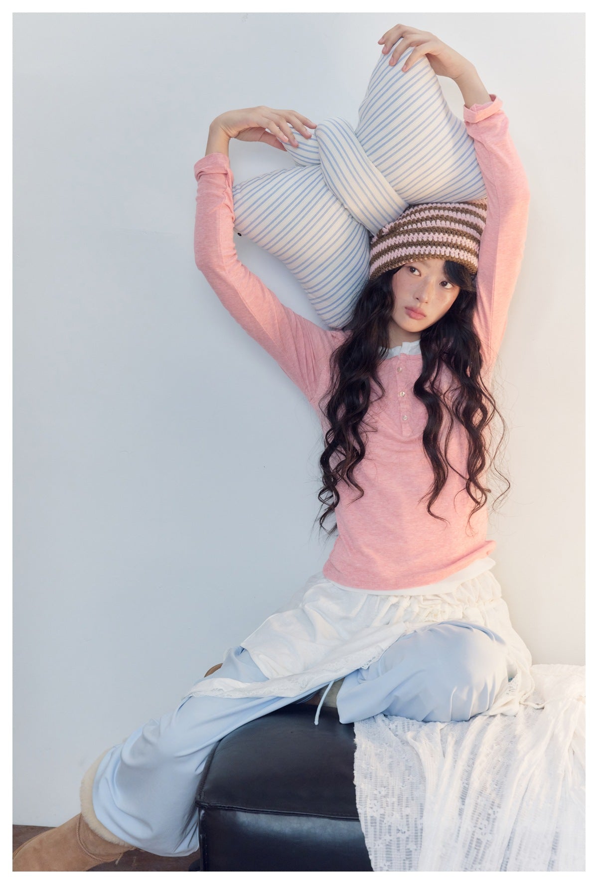 Model in a pink henley and pale blue pants holds striped pillows above her head, wearing a brown-and-pink knit hat.