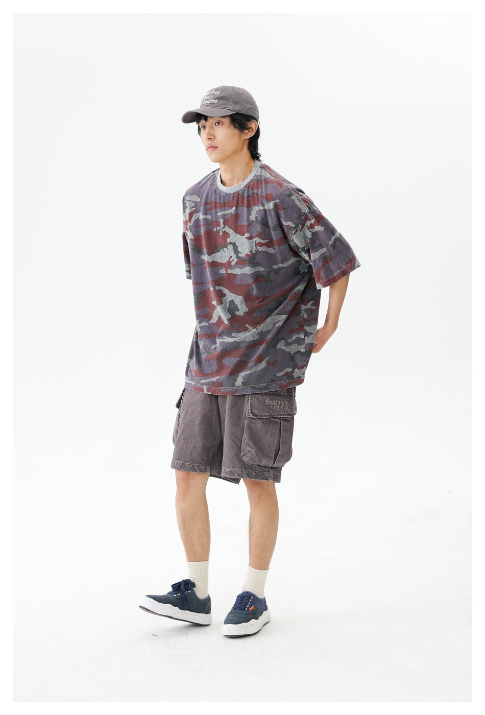 Man wearing an oversized camouflage T-shirt with a gray collar, brown cargo shorts, a gray cap and navy sneakers