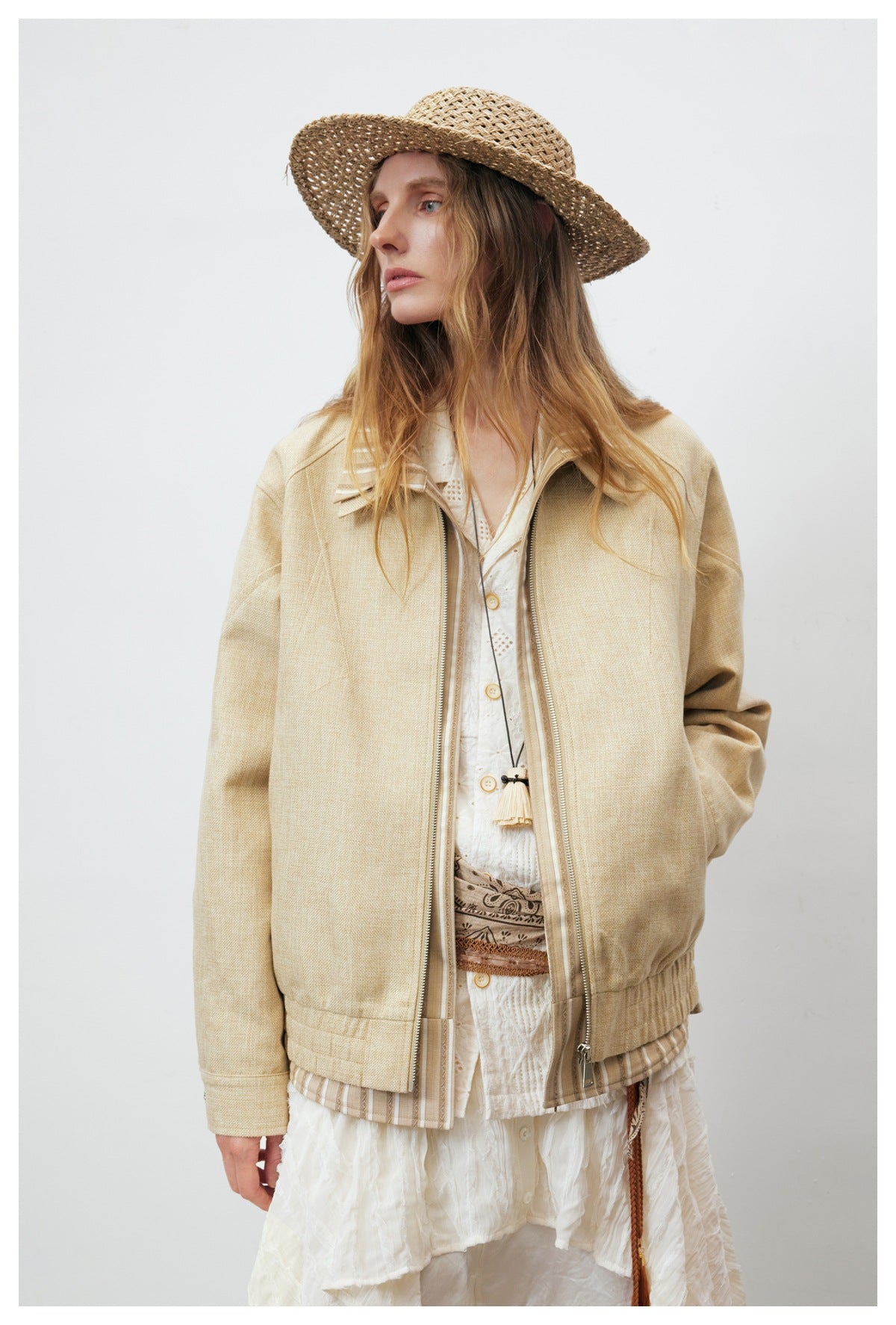 Model wearing open khaki zip jacket with striped trim over white layered outfit and straw hat