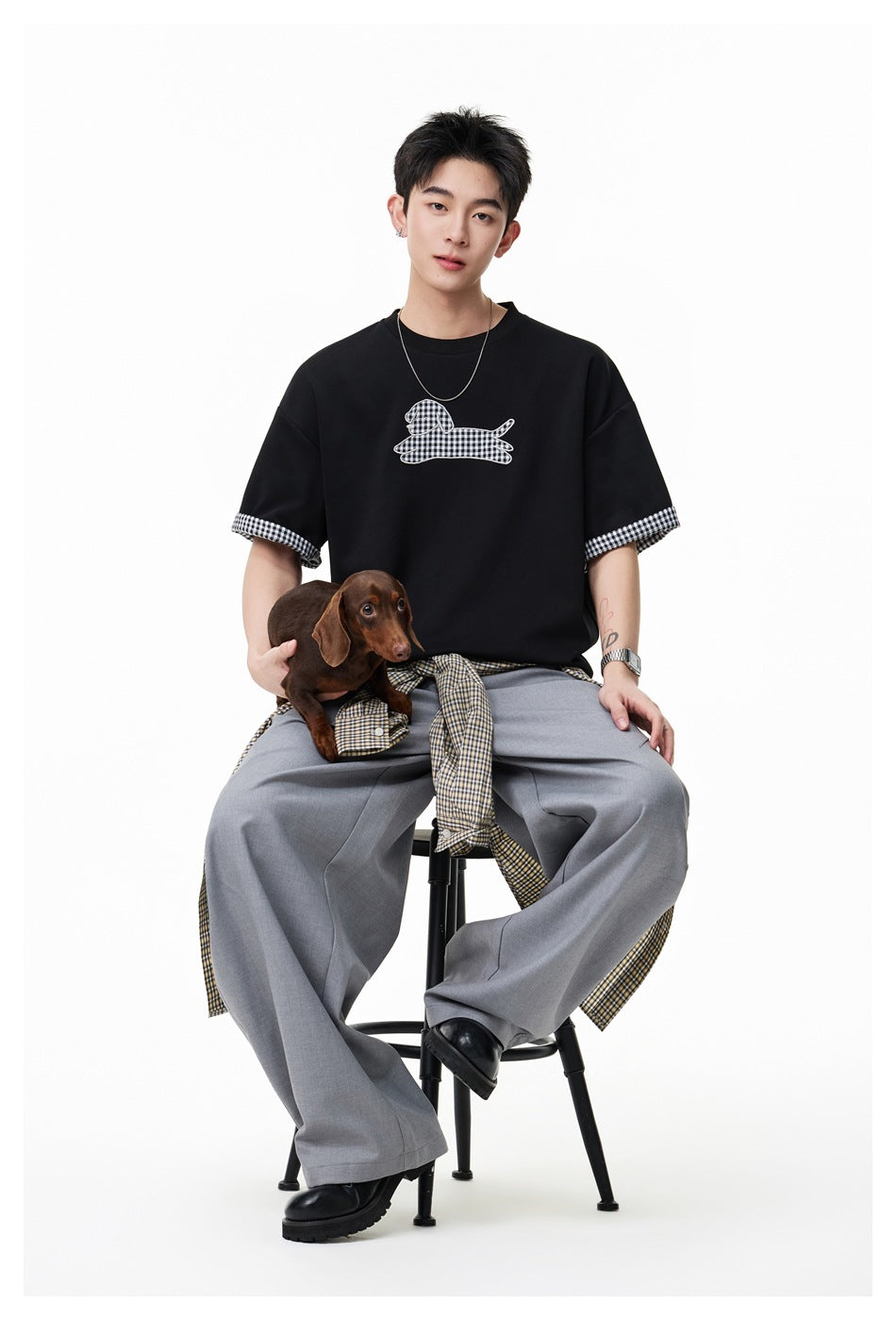 Man wearing a black dog-graphic T-shirt with checked sleeve cuffs and gray wide-leg trousers, seated with a dachshund.