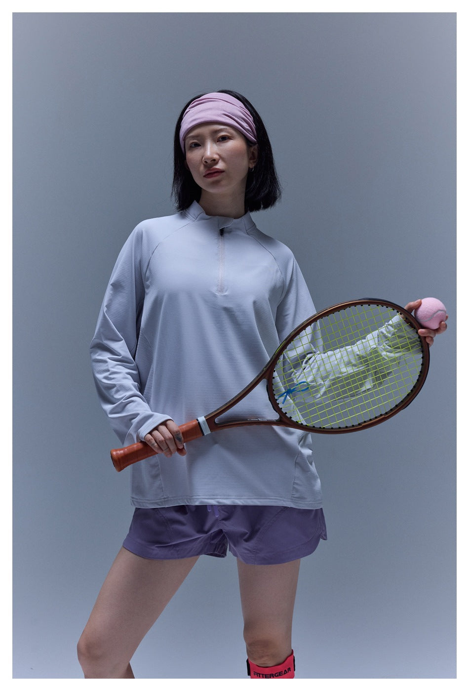 Model wearing a pale lavender-gray long-sleeve quarter-zip tennis top and purple shorts, holding a racket and ball.