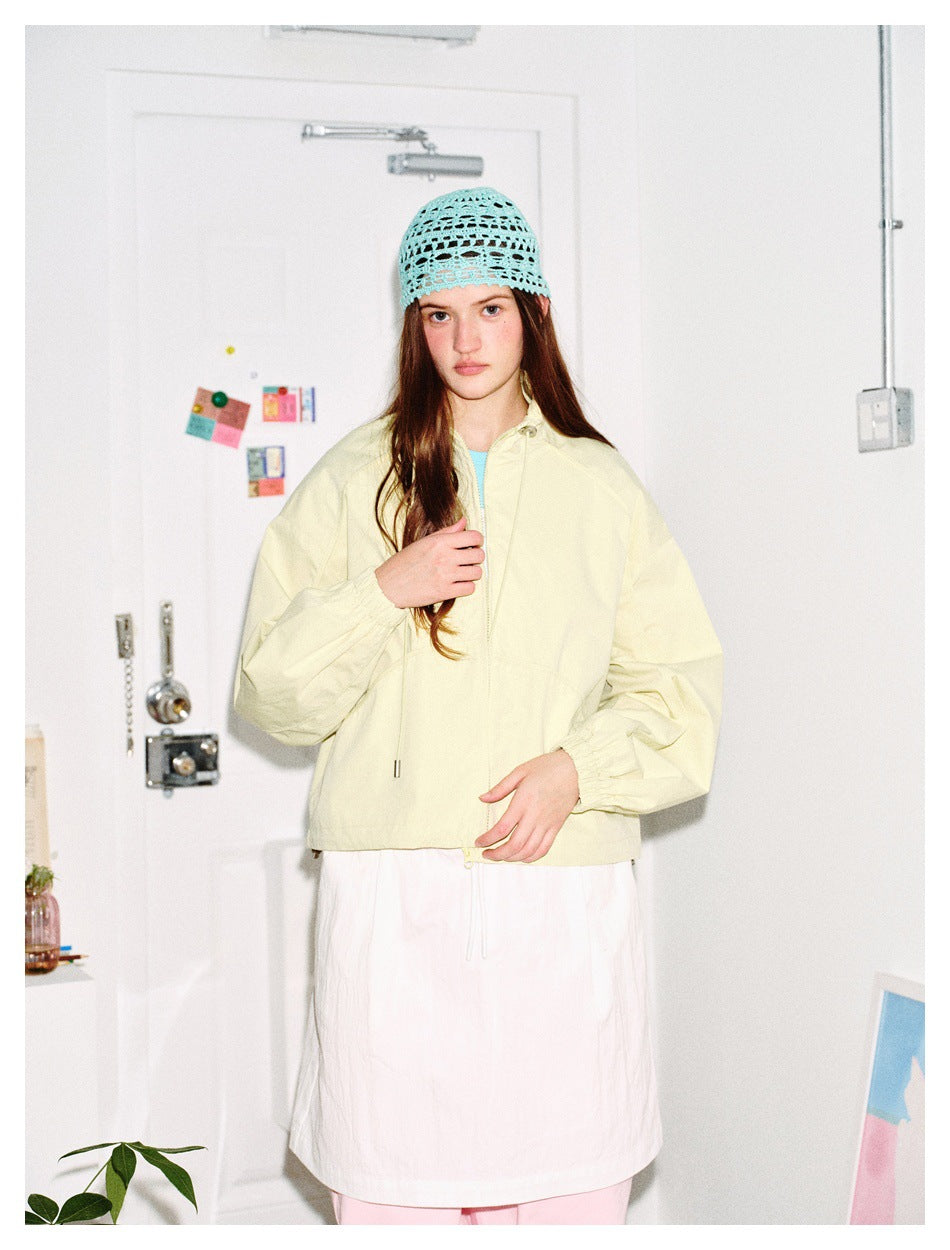 Model in light yellow cropped jacket with gathered cuffs, white skirt and light blue crochet hat, hand at chest