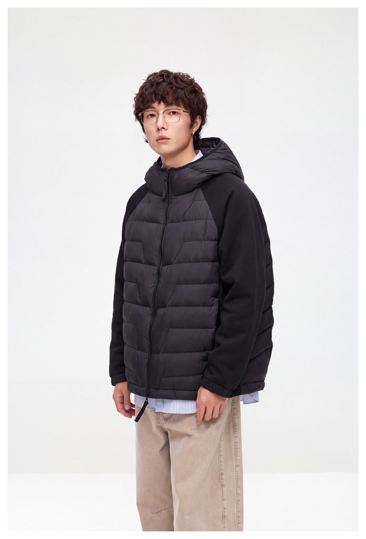 Model in a black hooded jacket with horizontal quilted front panels and solid sleeves, shown in a three-quarter view.