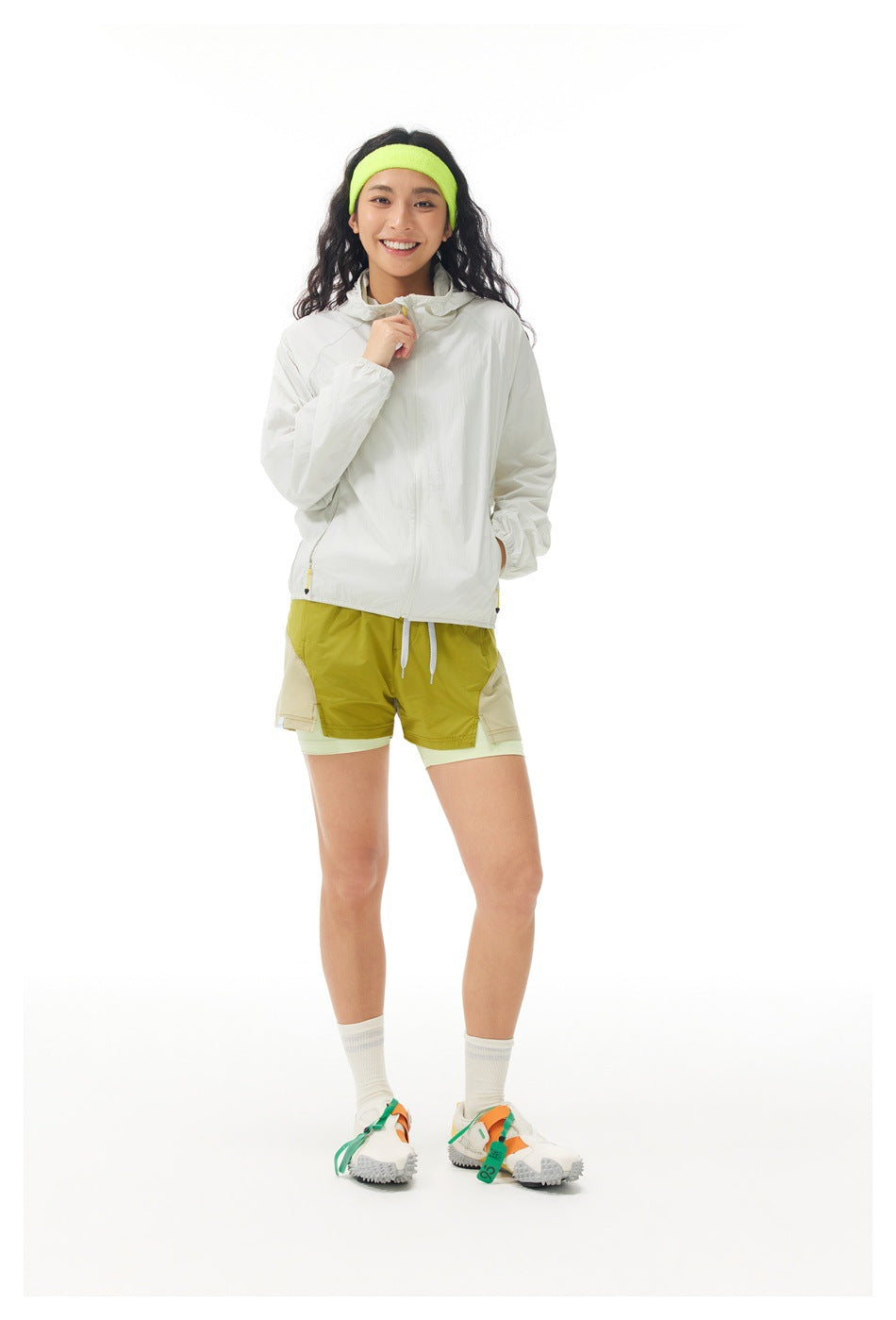 Full-length front view of a model wearing an off-white jacket, olive layered shorts, and a neon yellow-green headband.