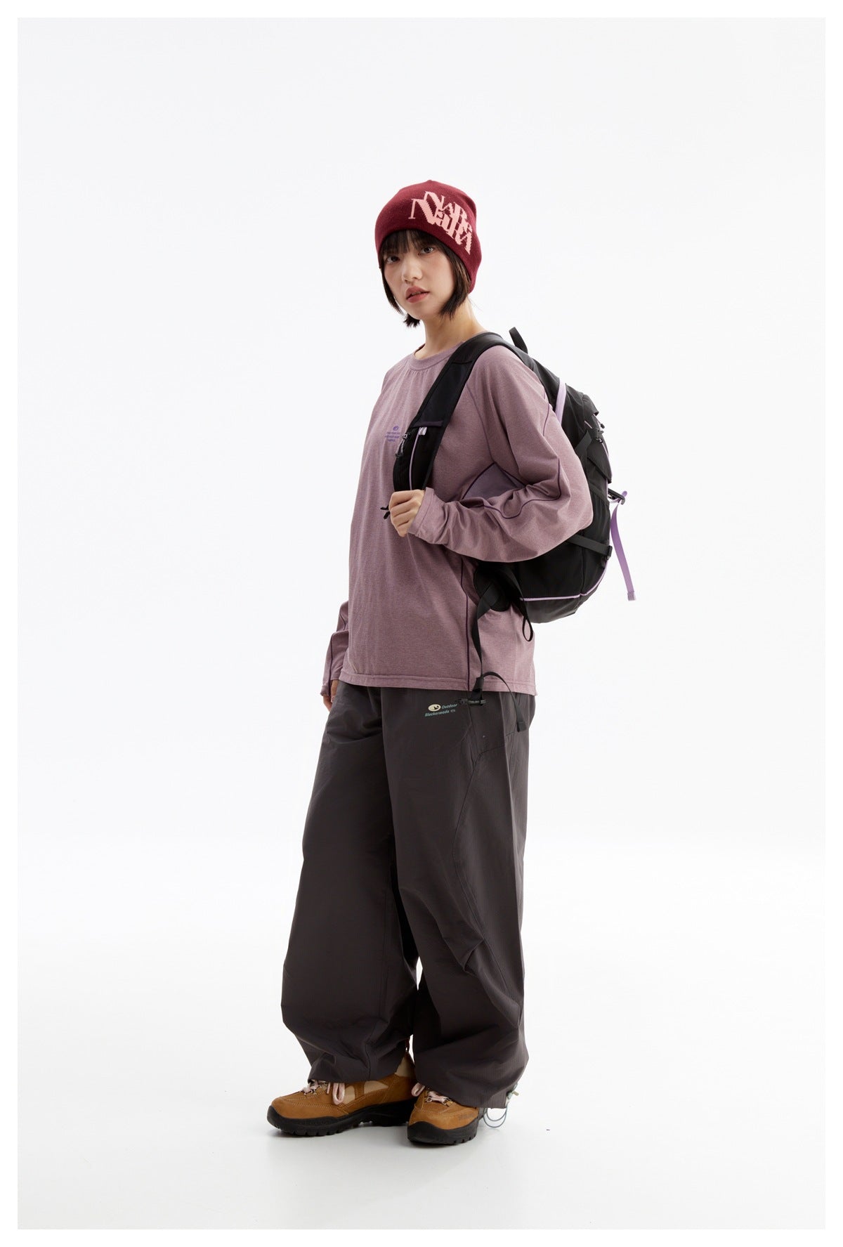 Model in a mauve long-sleeve top and loose dark pants, wearing a black backpack with lilac trim.