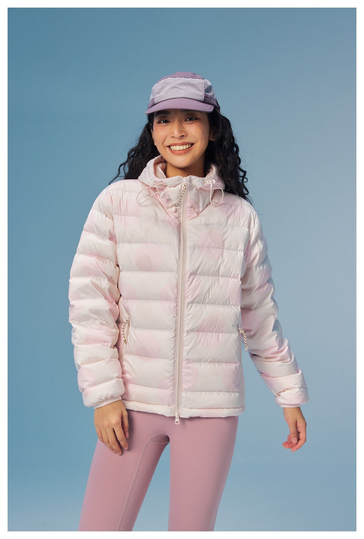 Model wearing a pale pink quilted hooded jacket with zip pockets and mauve leggings, shown from the front.