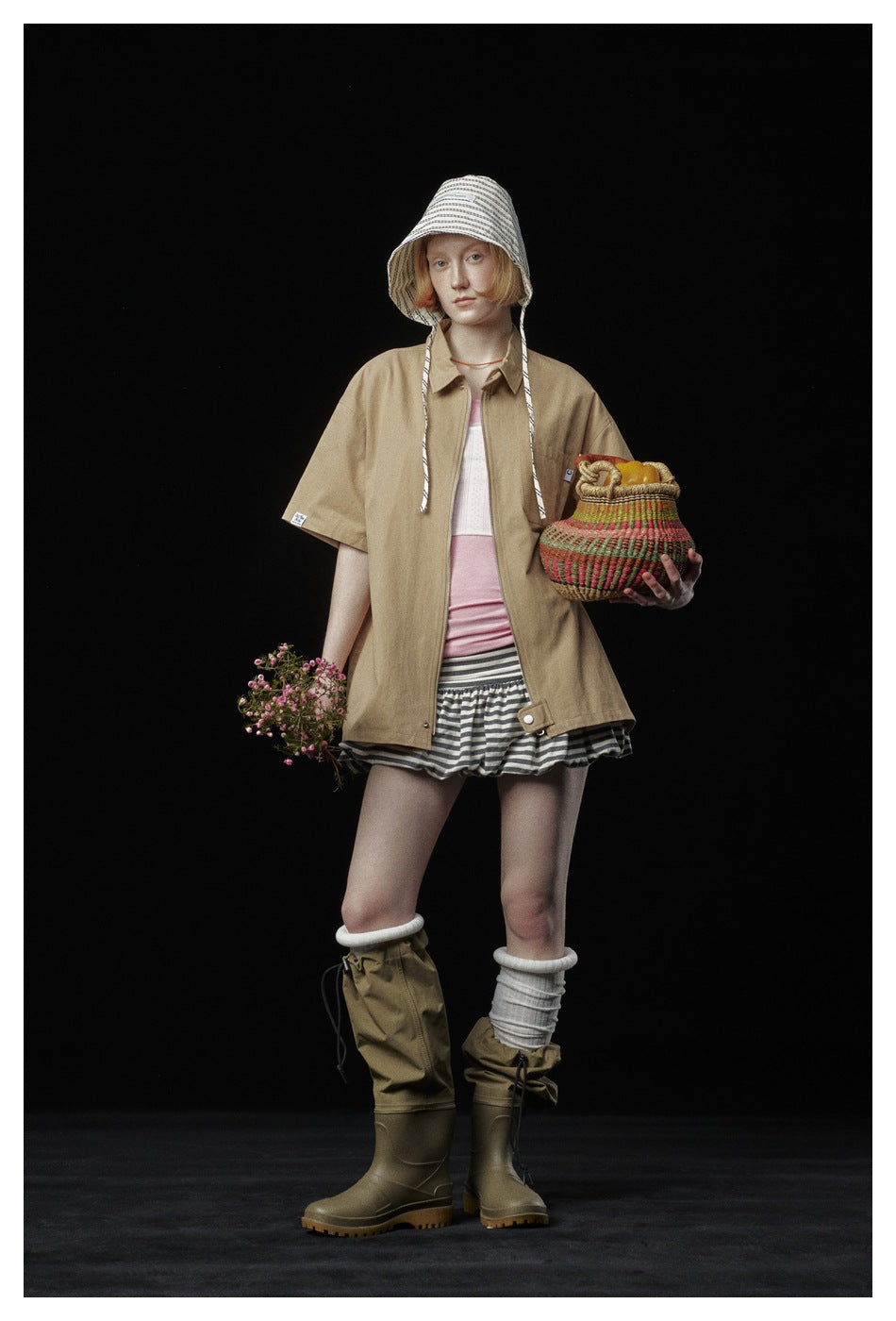 Model wearing a loose tan short-sleeve collared shirt over a pink top and striped shorts.