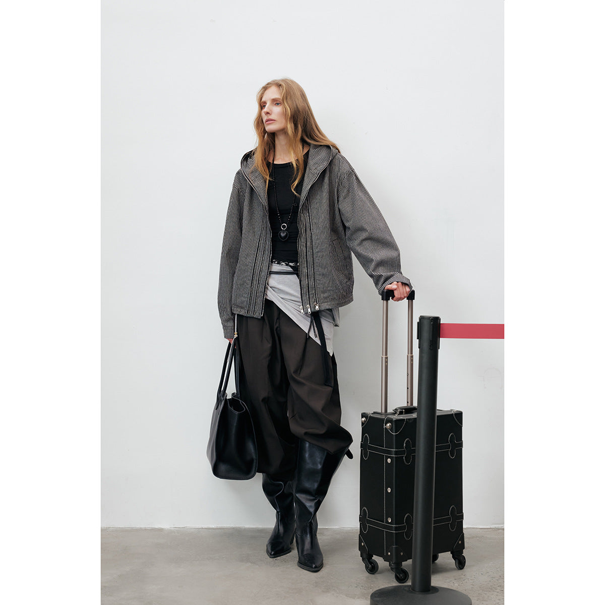 Full-length model wearing an open pinstriped hooded jacket, dark trousers and boots beside a suitcase.