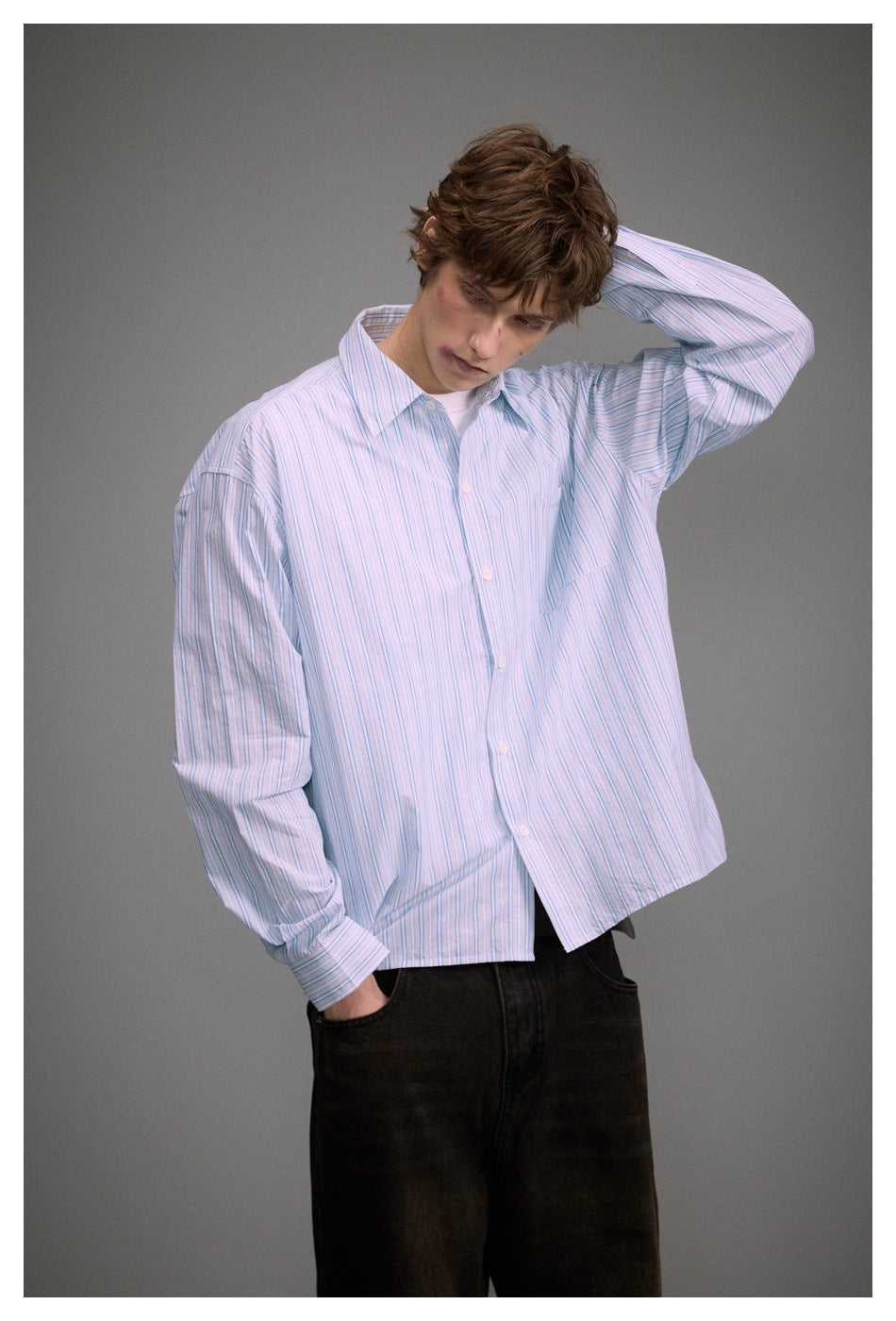 Model in a loose blue and white striped button-up shirt with one hand behind the head, over dark pants