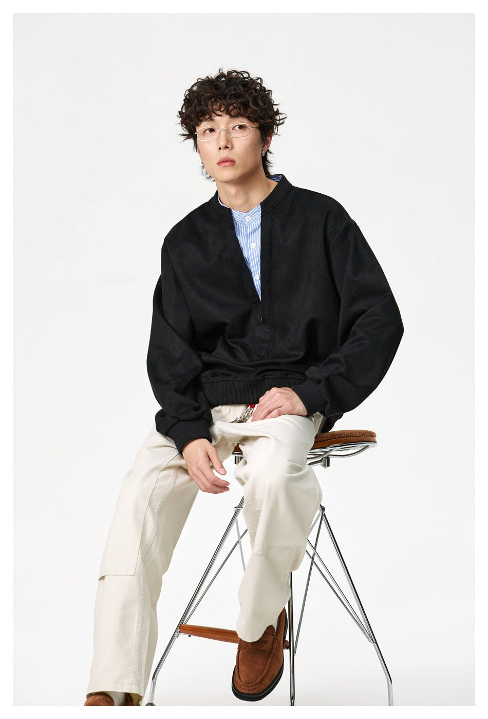 Seated model wearing a black oversized split-neck pullover over a blue striped shirt with cream trousers.