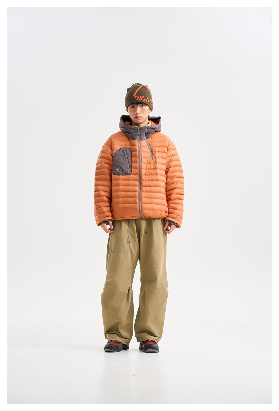 Front view of a model wearing an orange quilted hooded jacket with a gray chest pocket and loose olive trousers.
