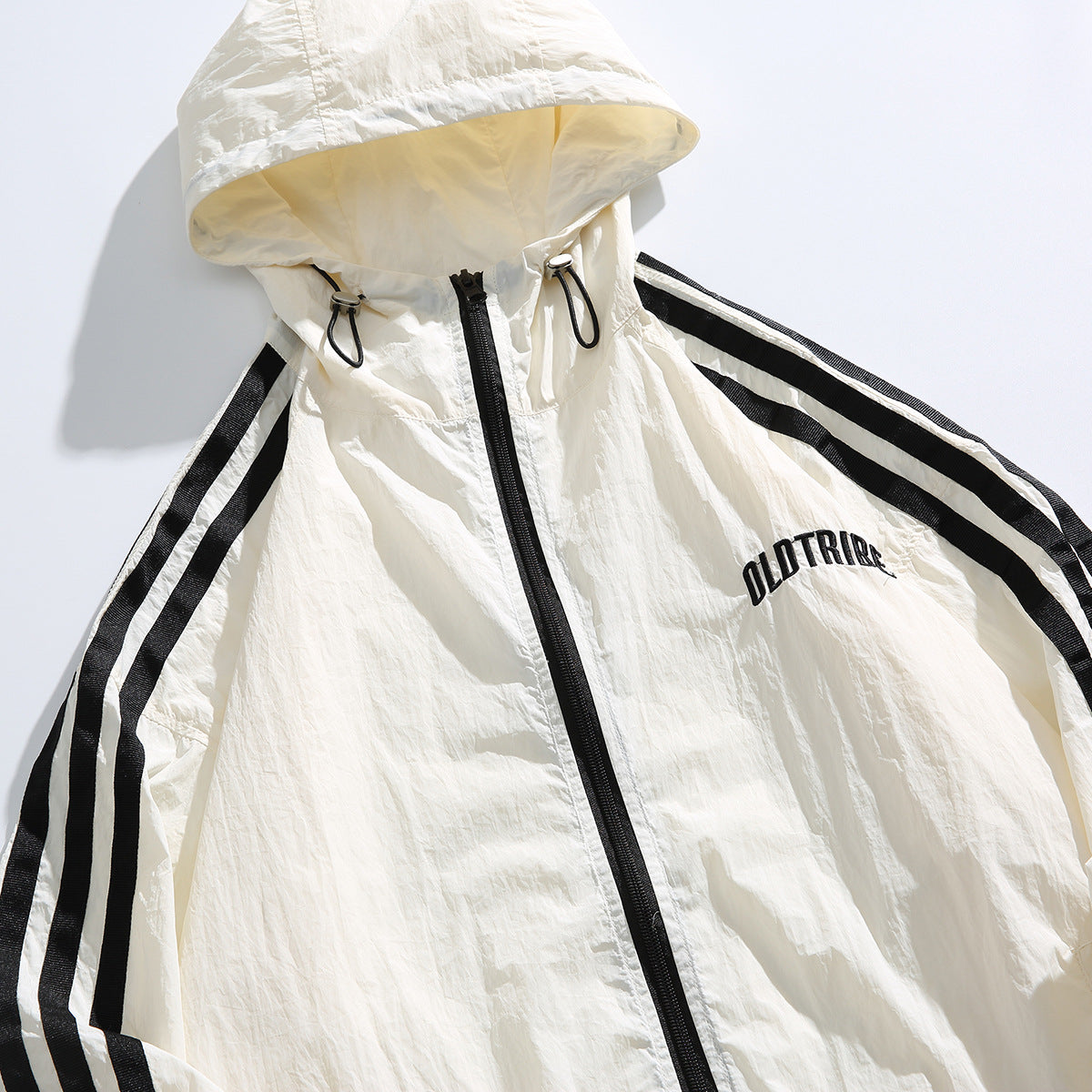 Close-up of an ivory hooded jacket with black sleeve stripes, a front zipper, drawcords, and embroidered chest lettering.