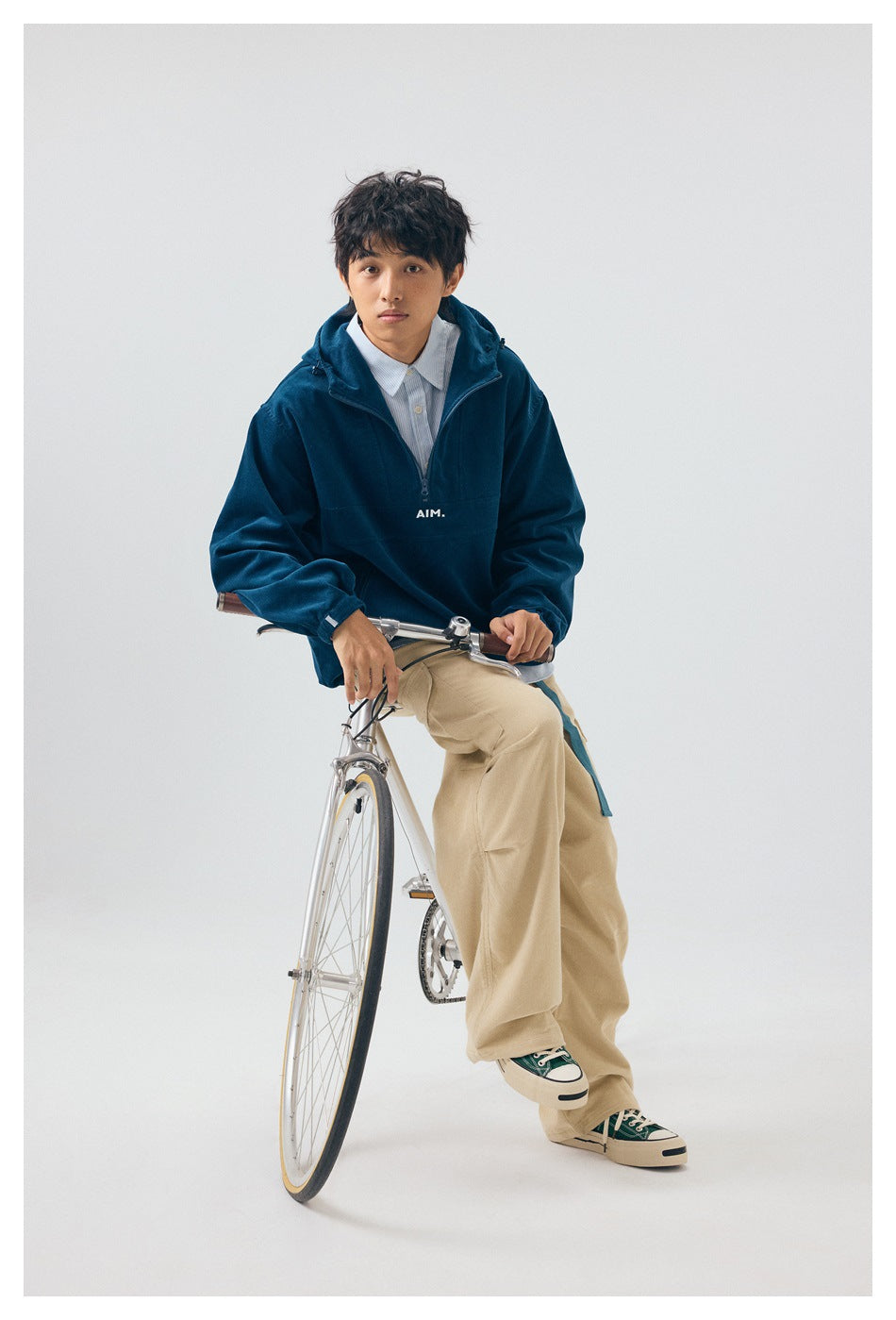 Model seated on a bicycle wearing a deep teal zip-neck hooded pullover with small white AIM lettering.
