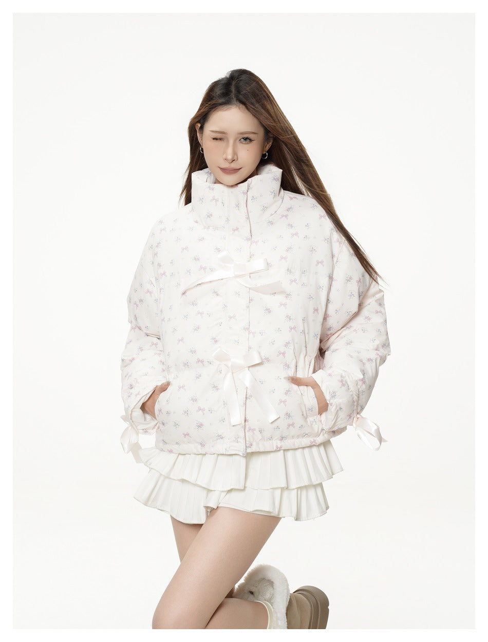 Model wearing a white cropped jacket with a high collar, pastel bow-and-floral print, satin ribbon ties, and a layered white
