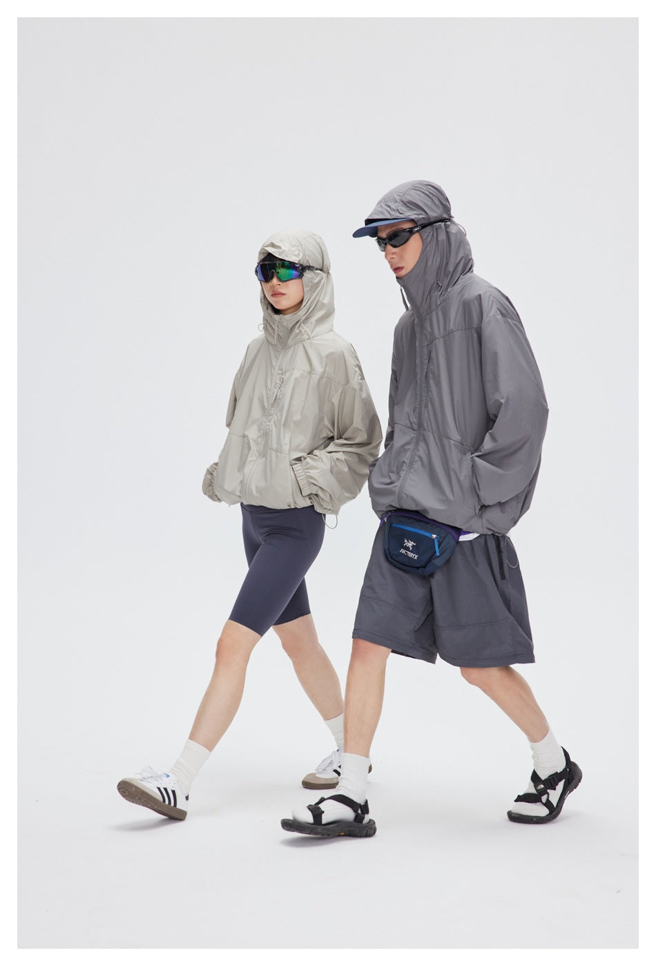 Two models walk in light beige and gray hooded windbreakers, with navy shorts or cycling shorts.