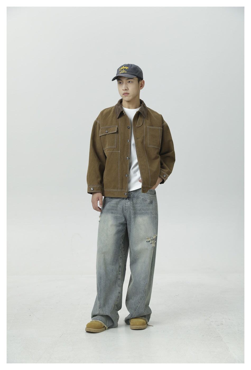 Man wearing olive brown work jacket over a white tee with light wash wide-leg jeans and a cap, front view
