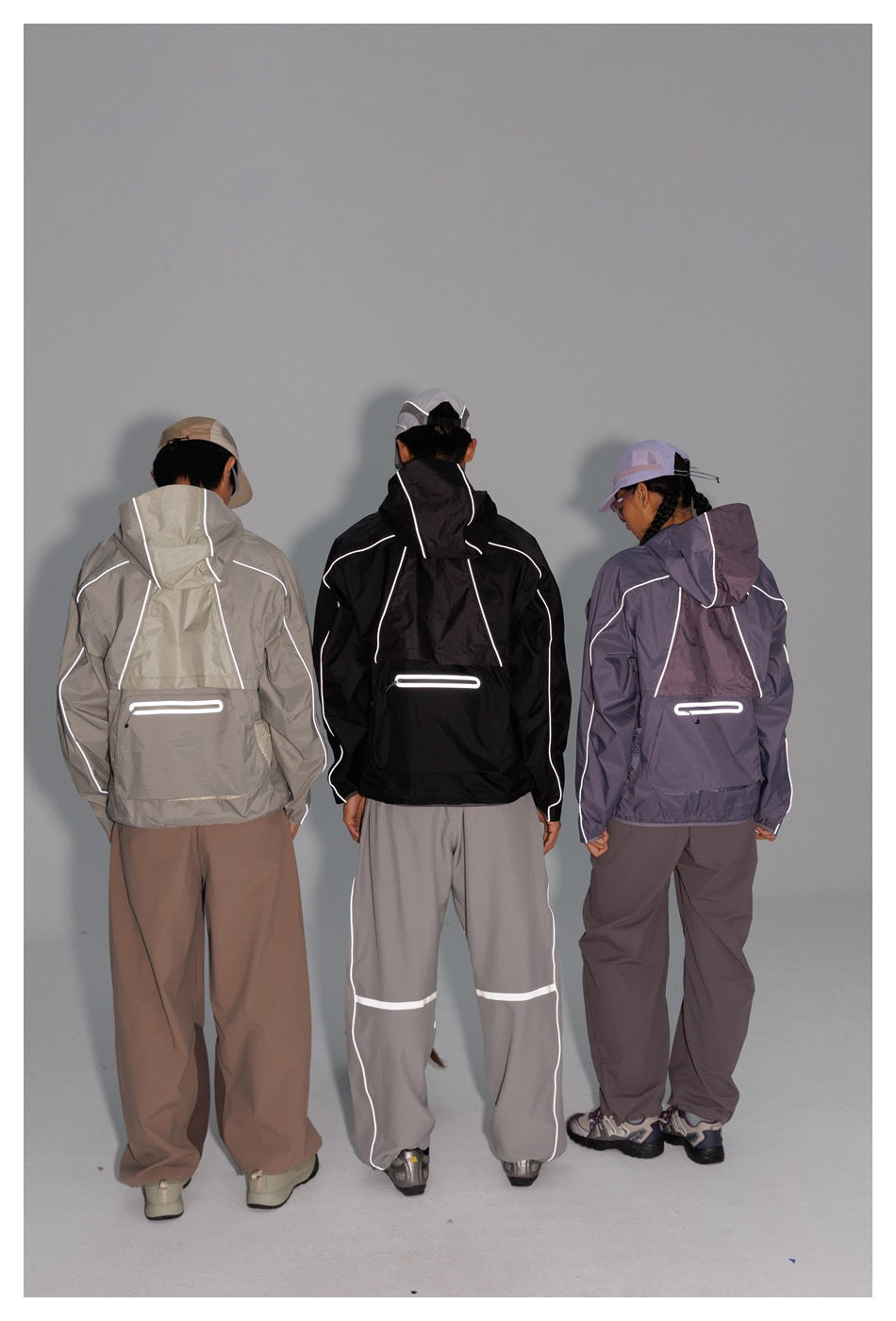 Back view of three models wearing light-gray, black, and lavender hooded jackets with reflective trim and rear pockets.