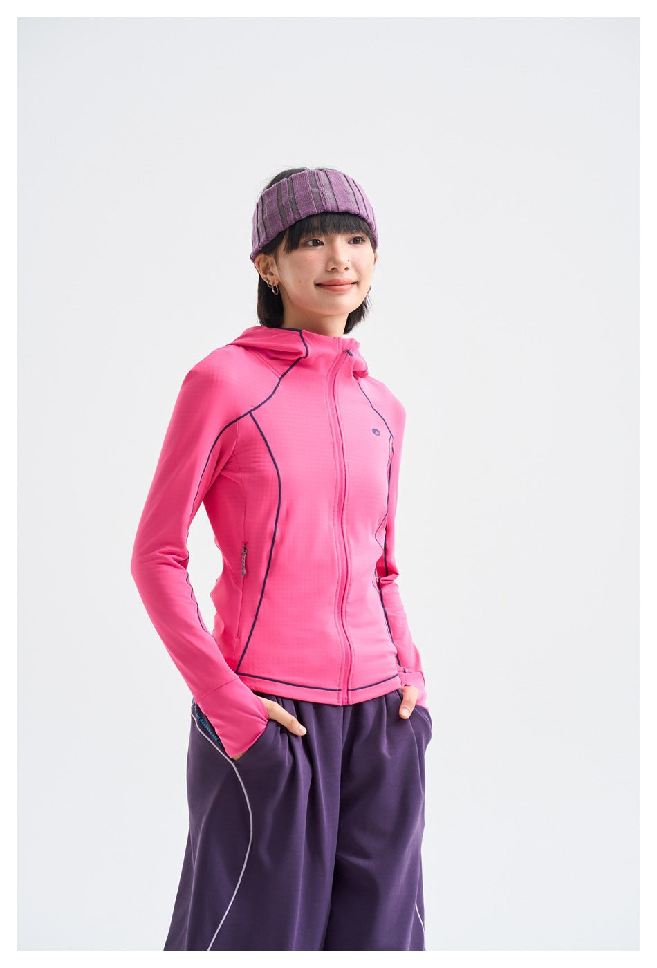 Three-quarter view of a model wearing a bright pink hooded zip jacket with dark piping and loose purple pants.