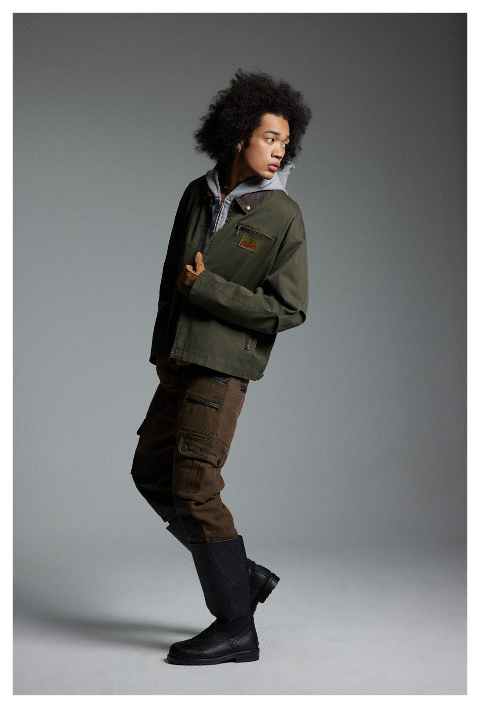 Model in a side pose wearing an olive work jacket, gray hoodie, brown cargo pants and black boots.