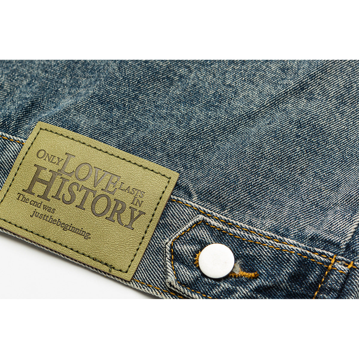 Close-up of a blue denim jacket’s waistband, metal button and sewn rectangular patch with English lettering.