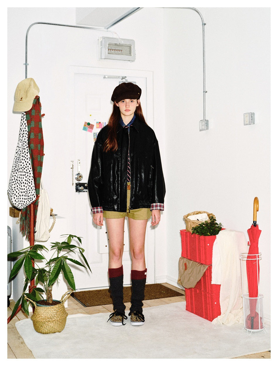 Model wearing a black leather-look jacket with plaid cuffs, olive shorts, a dark beret, and tall socks.
