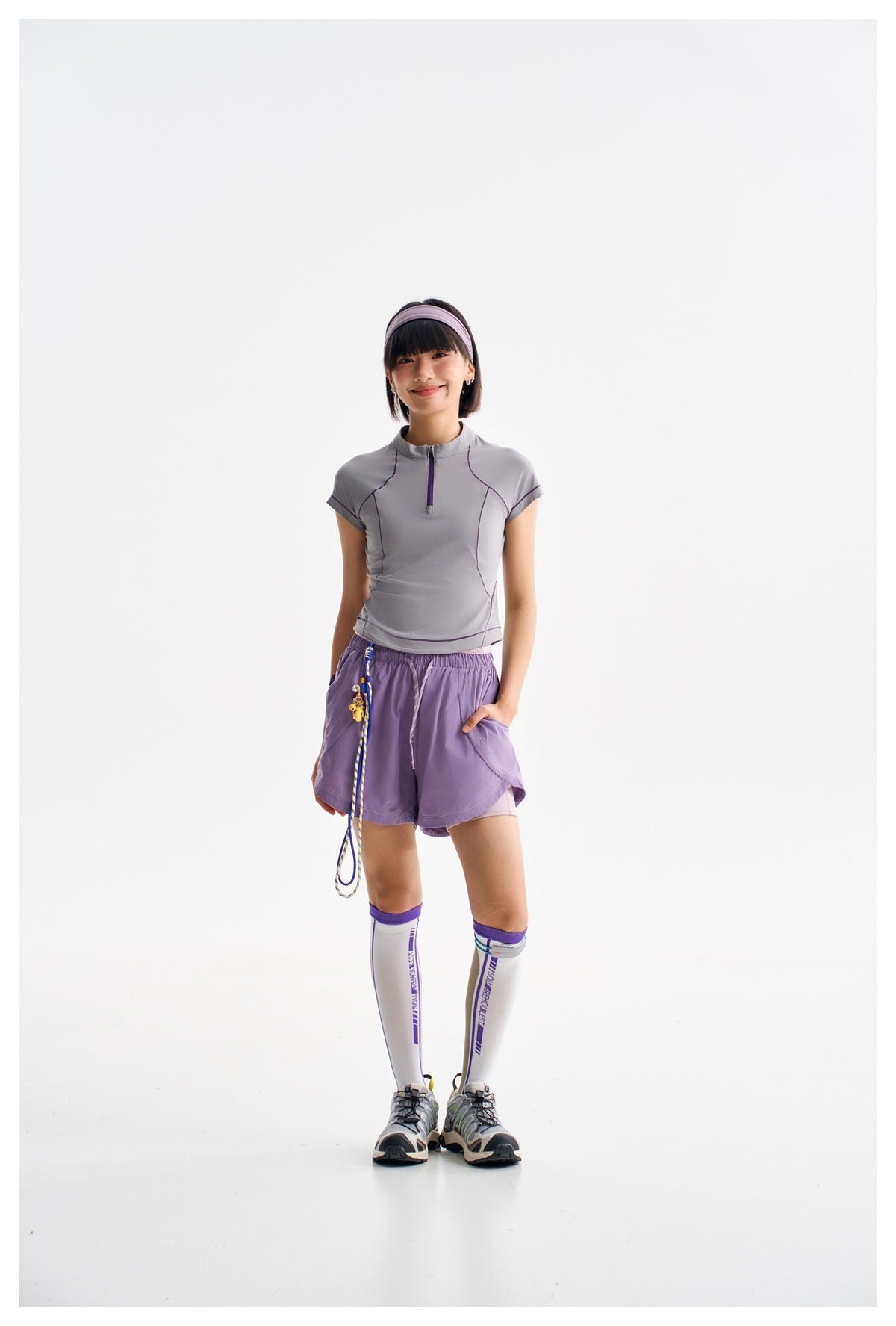 Model wearing a light gray zip-neck athletic top with purple piping and lavender shorts, posed against a white background.