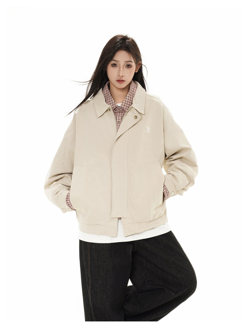 Model wearing a light beige collared jacket over a pink checked shirt and white top, with loose black jeans.