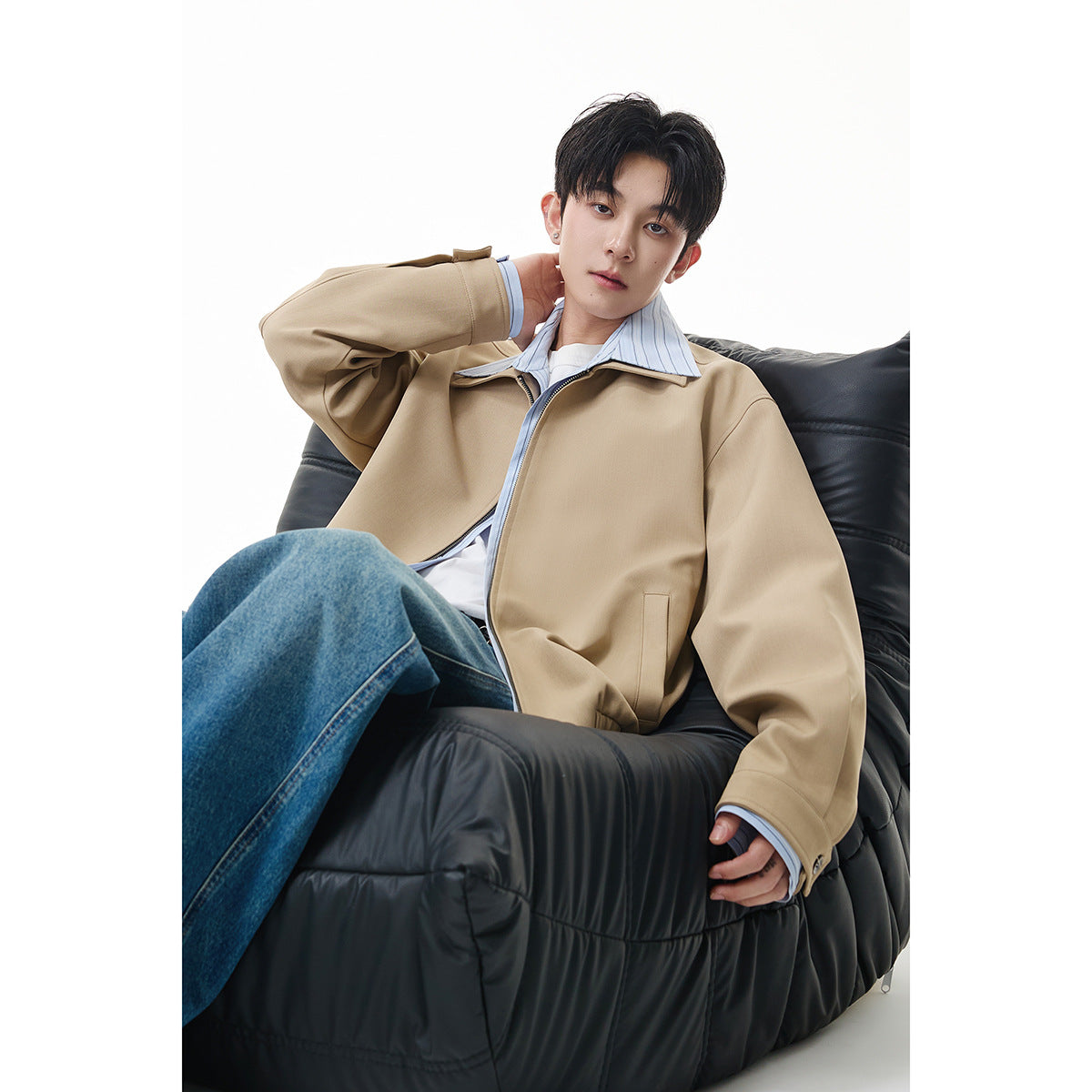 Model seated in a black chair wearing a relaxed beige zip-front jacket with a wide collar and blue jeans.