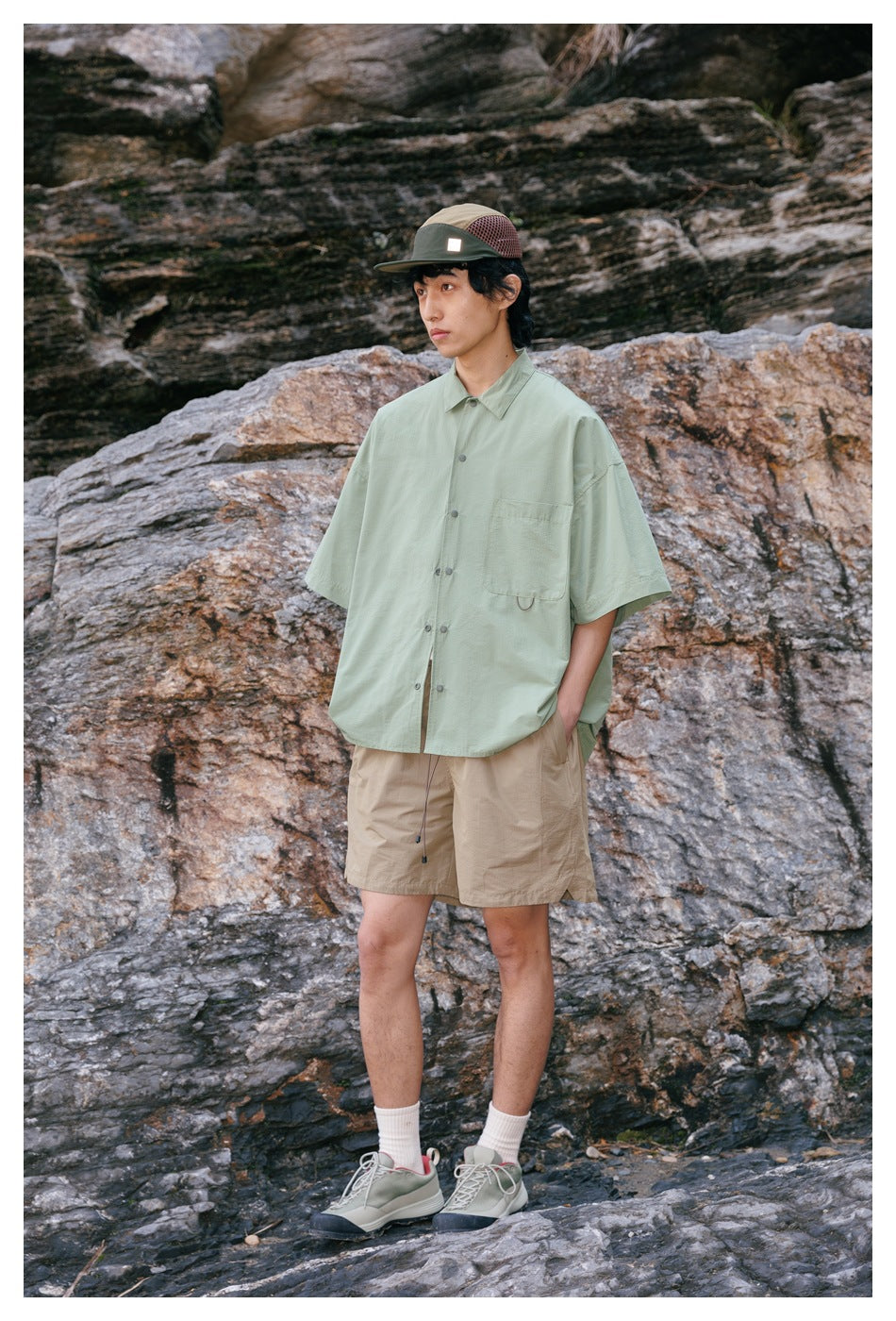 Side-view model wearing a pale sage-green short-sleeve button-up shirt and tan shorts against a rocky background.