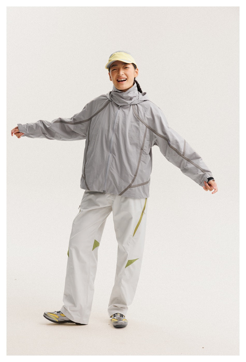 Model wearing a light gray hooded jacket and white pants with olive accents, arms extended to show the fit.